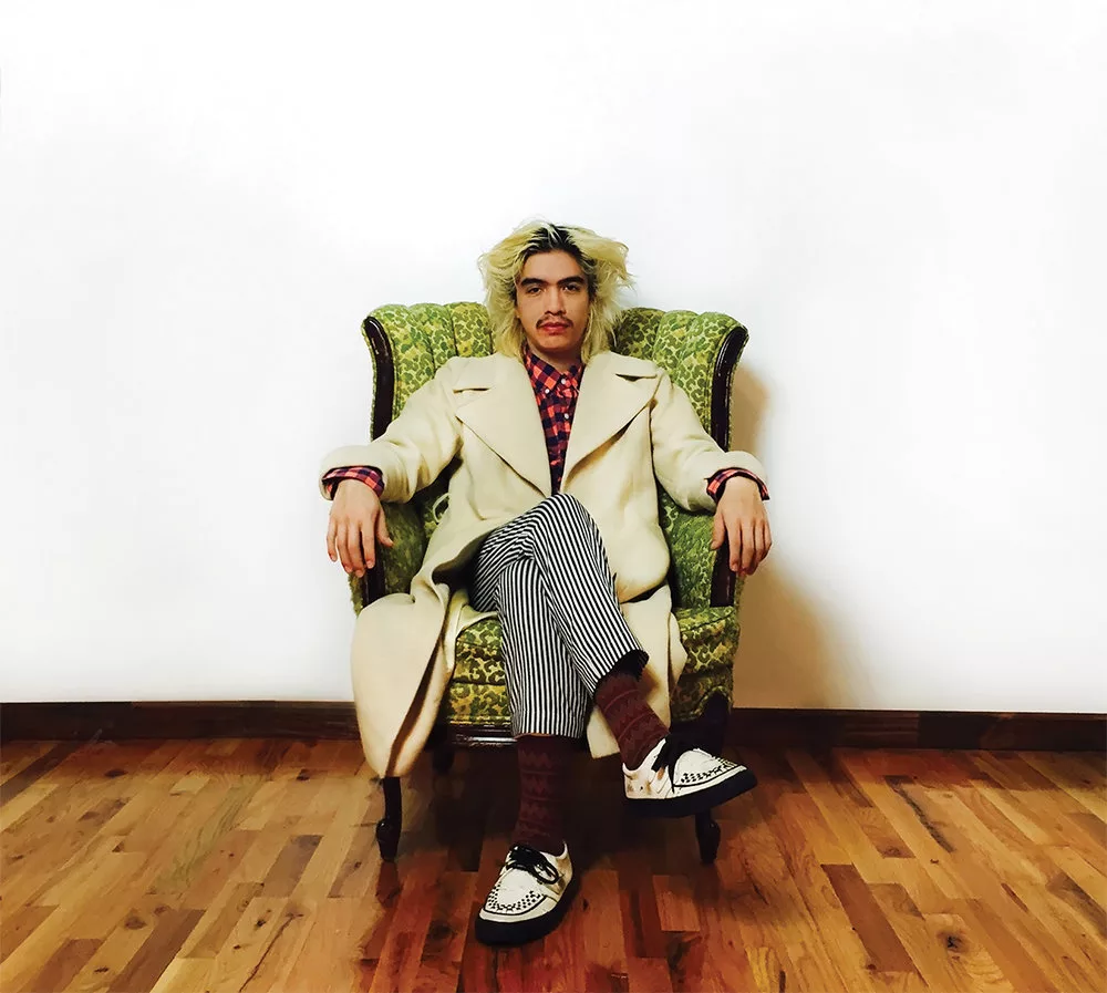 Album art for 'Evil,' depicting a photo portrait of the artist with platinum blond hair wearing a white coat, checkered shirt, striped pants, and black and white shoes sitting cross-legged in a green armchair in front of a white wall on light hardwood flooring